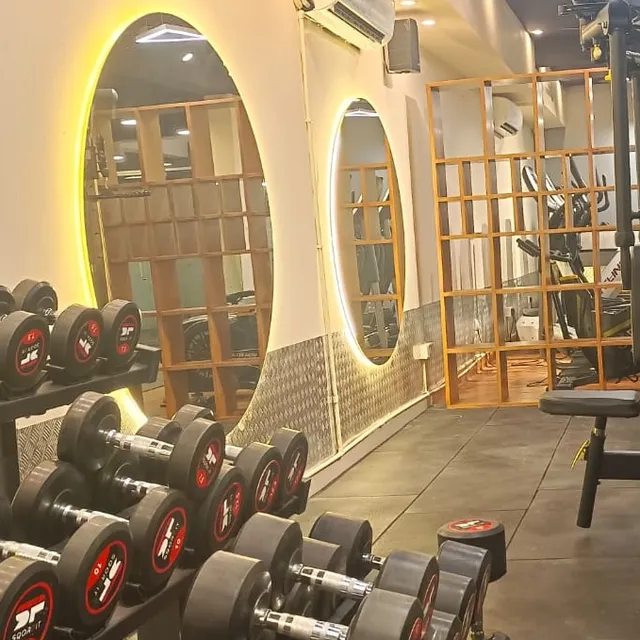A rack of dumbbells in the foreground, with two round backlit mirrors on the wall behind reflecting wooden cubby shelving and an air-conditioning unit.